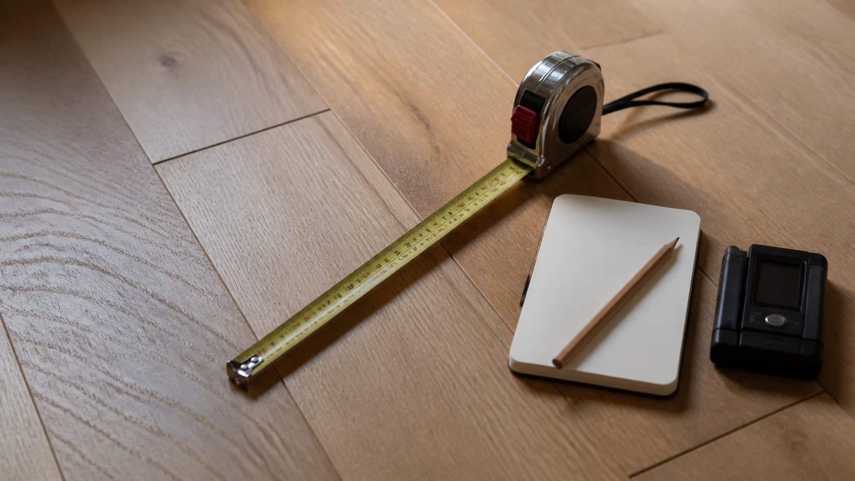 A steel tape measure run out across bare oak boards beside a moisture meter, a notebook and a carpenter pencil, on the floor of a Staten Island house